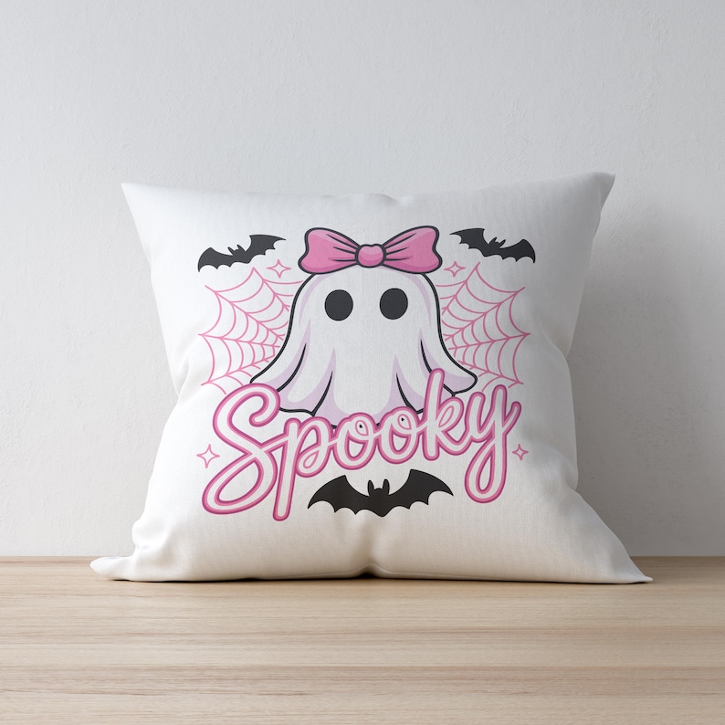 May include: White square pillow with a cartoon ghost wearing a pink bow. The word "Spooky" is written in pink script below the ghost, with bats and a spiderweb design. A Halloween-themed decorative pillow.