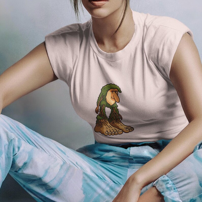 Op de afbeelding: Een wit t-shirt met een cartoon troll-achtig figuur met groen haar en bladachtige benen. Het wezen heeft een lange neus en staat op grote voeten. Het shirt wordt gecombineerd met een blauwe tie-dye broek.