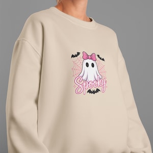 May include: Beige sweatshirt featuring a cartoon ghost with a pink bow, surrounded by bats and a spiderweb design. The word "Spooky" is written in pink script below the ghost. A fun, Halloween-themed design.