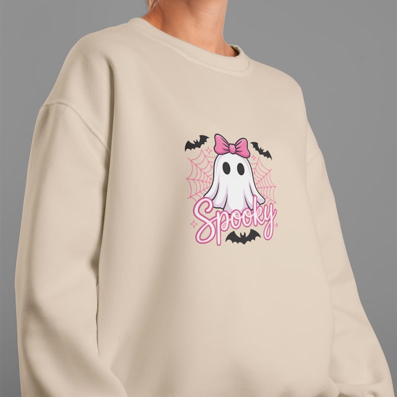 May include: Beige sweatshirt featuring a cartoon ghost with a pink bow, surrounded by bats and a spiderweb design. The word "Spooky" is written in pink script below the ghost. A fun, Halloween-themed design.