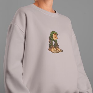 Op de afbeelding: Een lichtmauve sweatshirt met een cartoon trollenontwerp. De trol heeft een groene, bladerrijke hoed en baard, een lange neus en grote blote voeten. Het ontwerp is gecentreerd op de borst.