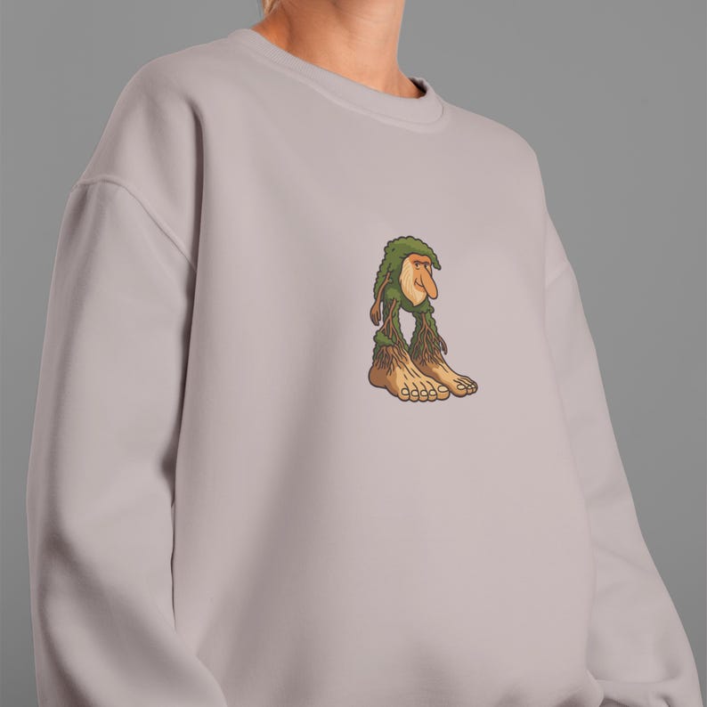 Op de afbeelding: Een lichtmauve sweatshirt met een cartoon trollenontwerp. De trol heeft een groene, bladerrijke hoed en baard, een lange neus en grote blote voeten. Het ontwerp is gecentreerd op de borst.