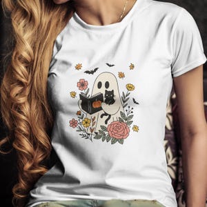 Puede incluir: Camiseta blanca con un fantasma de dibujos animados leyendo un libro y sosteniendo un gato negro. El fantasma est&aacute; rodeado de flores, hojas y murci&eacute;lagos. El libro tiene una calabaza en la portada. Una divertida camiseta gr&aacute;fica con tem&aacute;tica de Halloween.