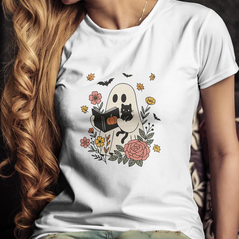 Puede incluir: Camiseta blanca con un fantasma de dibujos animados leyendo un libro y sosteniendo un gato negro. El fantasma est&aacute; rodeado de flores, hojas y murci&eacute;lagos. El libro tiene una calabaza en la portada. Una divertida camiseta gr&aacute;fica con tem&aacute;tica de Halloween.