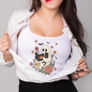 Puede incluir: Camiseta sin mangas blanca con un fantasma leyendo un libro, una calabaza y un gato negro. El dise&ntilde;o incluye flores, murci&eacute;lagos y hojas de oto&ntilde;o. La camiseta tiene cuello redondo.