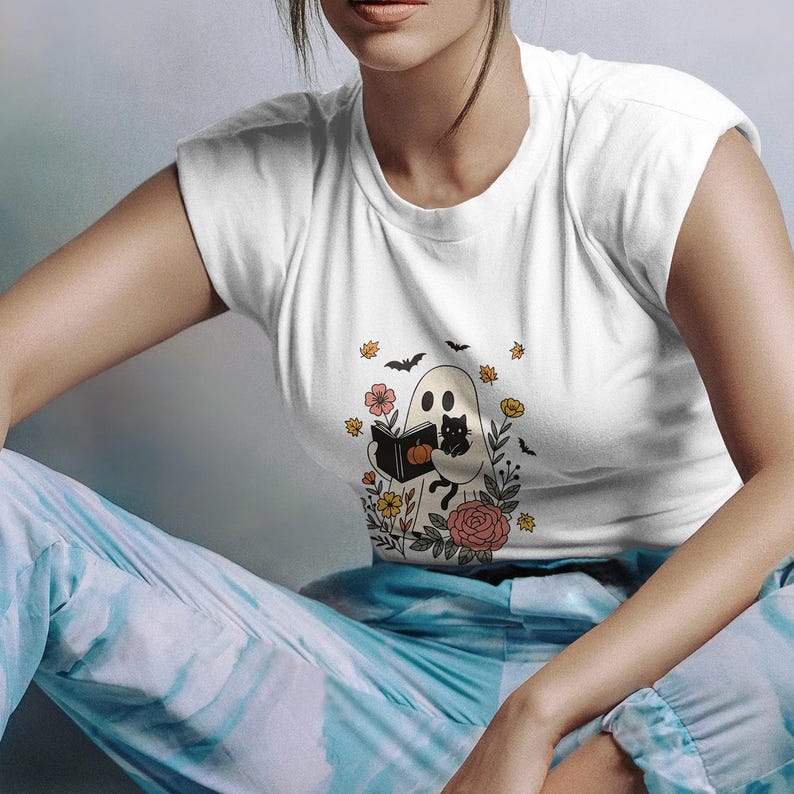 Puede incluir: Camiseta blanca con un dise&ntilde;o de Halloween. El gr&aacute;fico muestra un fantasma leyendo un libro, con un gato negro, flores, murci&eacute;lagos y una calabaza. La camiseta es de cuello redondo.