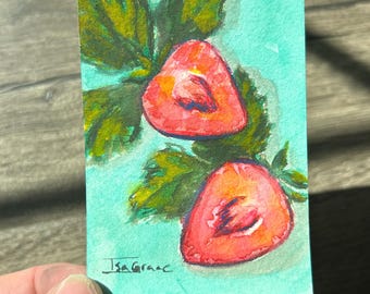 May include: A rectangular watercolor painting on paper featuring two sliced strawberries with green leaves against a light turquoise background. The artwork displays visible brushstrokes and the signature Isa Grace in the bottom left corner.