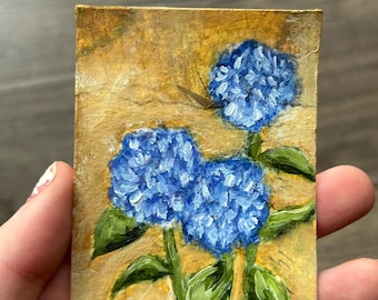 May include: A small rectangular painting on a textured surface featuring three vibrant blue hydrangea flower clusters with green leaves. The background is a warm, golden-yellow tone with visible brushstrokes throughout the entire composition.