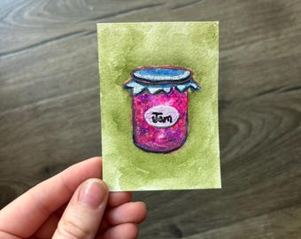 May include: A small rectangular painting on paper featuring a vibrant pink jar of jam with a blue lid. The jar has a white oval label with the word Jam written in black ink. The background is a textured olive green wash of watercolor paint.