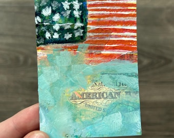 May include: A rectangular art card featuring a painted American flag in the upper section with white stars on a dark green field and red and white stripes. The lower section is painted in light blue with the text AMERICAN UNITED S visible.