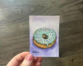 May include: A small rectangular watercolor painting featuring a golden-brown donut with light blue icing and dark blue sprinkles. The donut is set against a soft purple background, and the artist signature "J. Grace" is visible in the bottom left corner.