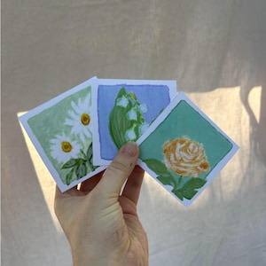May include: Three square watercolor paintings of flowers are held in a hand. The paintings feature a daisy, lily of the valley, and a rose. Each painting has a colored background: green, blue, and teal. The paintings are on white paper.