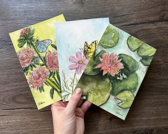 May include: Three rectangular art prints featuring nature illustrations. The left print shows pink flowers and a blue butterfly on a yellow background. The centre print depicts a yellow butterfly on a pink flower. The right print shows a water lily.