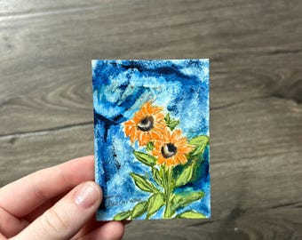 May include: A small rectangular painting on paper featuring two orange sunflowers with dark centres and green leaves. The background is a textured, deep blue wash with visible brushstrokes. A signature is visible in the bottom left corner.