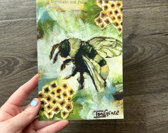 May include: A rectangular art print featuring an oil painting of a bumblebee centred between two honeycomb patterns. The background is a textured mix of light green and blue brushstrokes. The text Moonlight and Fairie is at the top, with the signature IsaGrace at the bottom right.