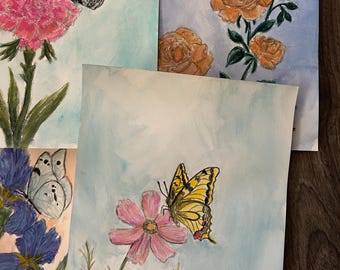 May include: A collection of four watercolour paintings on paper featuring botanical and insect themes. The artworks display a pink carnation with a black butterfly, orange roses with a bumblebee, blue irises with a white butterfly, and a pink cosmos with a yellow swallowtail butterfly. Each piece features delicate brushwork and soft colour washes.