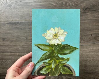 May include: A painting of a white daisy with a yellow center, green leaves, and a gray pot against a turquoise background. The artwork is held by a hand, showcasing the floral still life.