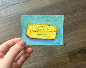 May include: A small rectangular watercolor painting on paper depicting a yellow stick of butter against a blue background. The butter is labeled with the text Grade A, Unsalted, BUTTER, and Net Wt. 4 oz (113g) written in black ink.