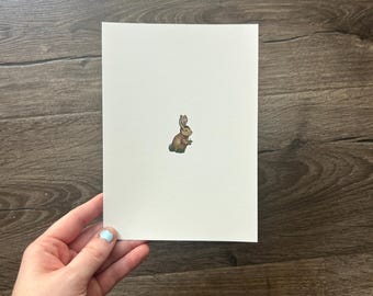 May include: A rectangular white card featuring a small, centred illustration of a brown rabbit sitting on a patch of green grass. The rabbit is depicted in profile, facing towards the right side of the card, with its ears upright and alert.