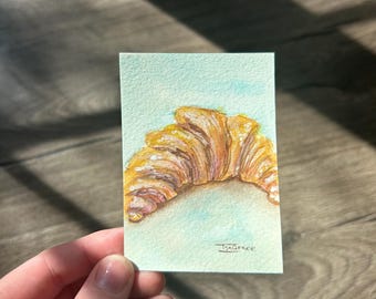 May include: A small rectangular watercolor painting on paper featuring a golden-brown croissant with layered, flaky textures against a light blue background. The artwork is signed with the text IsaGrace in the bottom right corner.