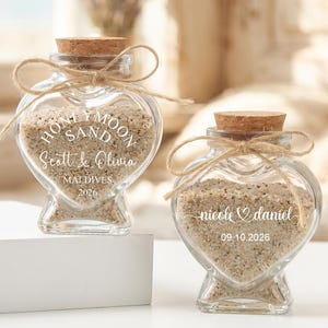May include: Two heart-shaped glass bottles filled with sand, each sealed with a cork and tied with twine. One reads "Honeymoon Sand, Scott & Olivia, Maldives 2026." The other reads "nicole & daniel, 09.10.2026."