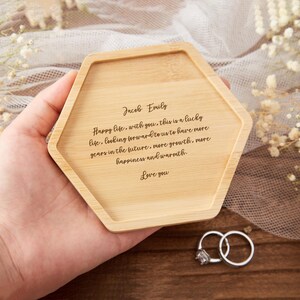 Op de afbeelding: Een lichtgekleurde, zeshoekige bamboe tray met gegraveerde tekst. De tekst luidt: "Jacob Emily Happy life, with you, this is a lucky life...Love you." Twee zilveren ringen met een diamant liggen naast de tray.