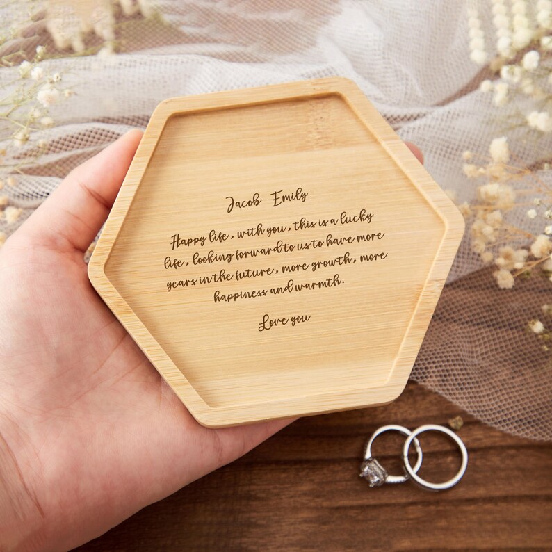 Op de afbeelding: Een lichtgekleurde, zeshoekige bamboe tray met gegraveerde tekst. De tekst luidt: "Jacob Emily Happy life, with you, this is a lucky life...Love you." Twee zilveren ringen met een diamant liggen naast de tray.