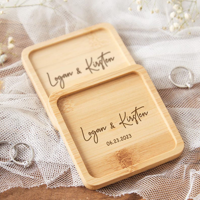 Op de afbeelding: Twee vierkante, lichtgekleurde houten ringendoosjes met afgeronde randen. Op het bovenste doosje staan de namen "Logan & Kirsten" gegraveerd in een sierlijk lettertype. Op het onderste doosje staan ook de namen en de datum "06.23.2023". Trouwringdoosjes.