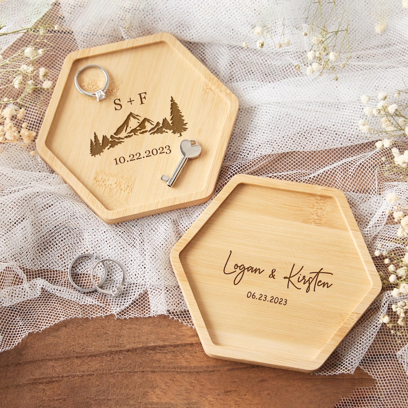 Op de afbeelding: Twee zeshoekige houten ringhouders. E&eacute;n heeft "S + F", berg- en boomontwerpen en "10.22.2023" gegraveerd. De andere heeft "Logan & Kirsten" en "06.23.2023" gegraveerd. Een sleutel en ringen zijn ook aanwezig.