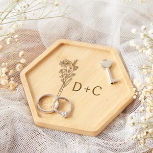 Op de afbeelding: Een houten, zeshoekige ringhouder met gegraveerd bloemmotief en initialen "D+C". Twee zilveren ringen en een kleine sleutel rusten op de houder. De achtergrond heeft witte stof en delicate witte bloemen.