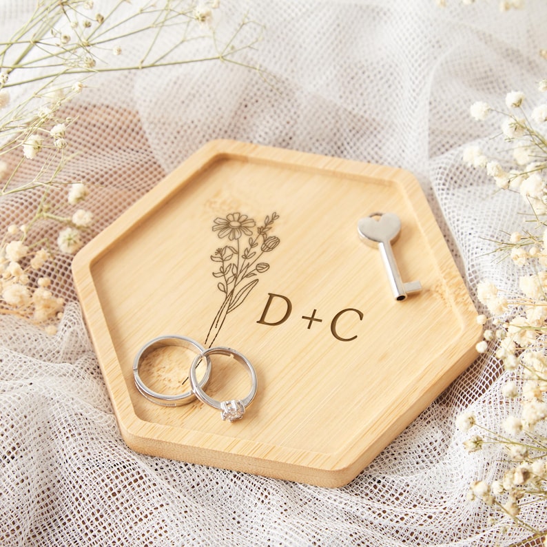 Op de afbeelding: Een houten, zeshoekige ringhouder met gegraveerd bloemmotief en initialen "D+C". Twee zilveren ringen en een kleine sleutel rusten op de houder. De achtergrond heeft witte stof en delicate witte bloemen.
