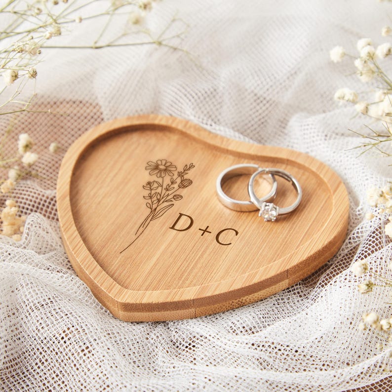 Op de afbeelding: Een hartvormige houten ringschaal met gegraveerd bloemmotief en de letters "D + C". Twee zilveren ringen liggen op de schaal. De schaal staat op een wit, getextureerd oppervlak.