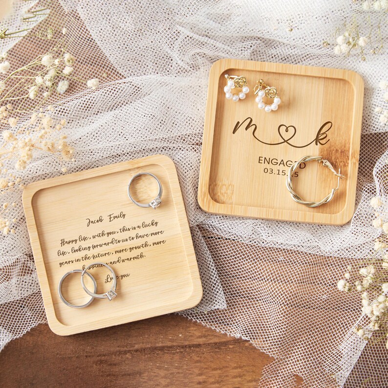 Op de afbeelding: Twee vierkante bamboe trays met gegraveerde tekst en sieraden. E&eacute;n tray heeft ringen, de andere heeft oorbellen en een oorring. Tekst op de trays bevat namen, data en een boodschap over een gelukkig leven.