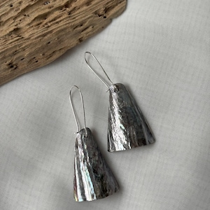 May include: A pair of silver-toned earrings with a textured, cone-shaped design. The earrings have a hammered metal appearance and are suspended from simple wire hooks. The background is a light, neutral fabric.
