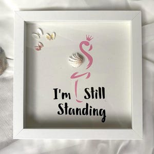 May include: White framed art with a pink flamingo design and the text "I'm Still Standing". The artwork includes seashells and small decorative elements. The overall theme is coastal and inspirational.