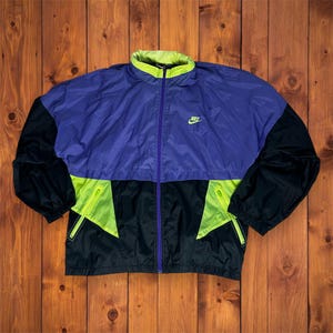 Könnte beinhalten: Vintage Nike Windjacke in Lila, Schwarz und Neongrün. Die Jacke hat einen durchgehenden Reißverschluss, einen hohen Kragen und das Nike-Logo auf der Brust. Die Ärmel und der untere Teil sind schwarz, mit neongrünen Akzenten.