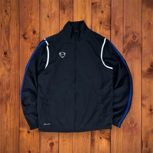 Könnte beinhalten: Schwarze Nike Trainingsjacke mit durchgehendem Reißverschluss. Sie hat weiße Akzente an den Schultern und blaue Streifen an den Ärmeln. Kleines Nike-Logo auf der Brust. Die Jacke ist aus leichtem Material. Der Text "DRI-FIT" ist sichtbar.