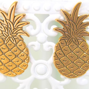 Op de afbeelding: Een paar gouden ananas oorbellen. De ananas zijn gedetailleerd met een getextureerd oppervlak en een reliëfdesign.