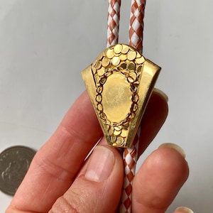 Puede incluir: Una corbata bolo dorada con un diseño geométrico. La corbata presenta un colgante decorativo con una superficie texturizada y un centro ovalado. El cordón trenzado es blanco y marrón. La corbata bolo es un accesorio de estilo occidental.