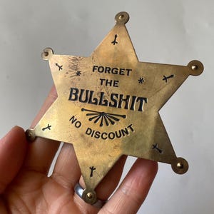 Könnte beinhalten: Ein messingfarbener Sheriff-Abzeichen im Vintage-Stil mit den Worten "FORGET THE BULLSHIT" und "NO DISCOUNT" in schwarzer Schrift. Das sternförmige Abzeichen hat dekorative Nieten und kleine Stern- und Kreuzdetails.