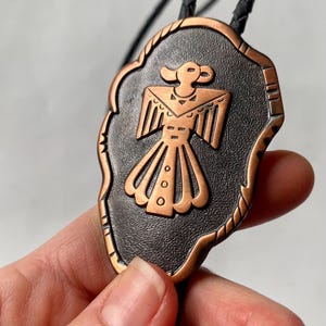 May include: A bolo tie with a copper-coloured Thunderbird design on a black background. The pendant has a decorative edge and is suspended from a braided black cord.