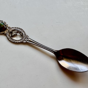 May include: Silver souvenir spoon with an ornate handle and a detailed bowl. The handle has a decorative design with a small, colourful inset and a cut-out shape. The spoon is likely made of metal.