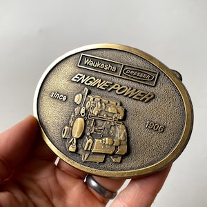 May include: Vintage brass belt buckle with an oval shape. The buckle features the words "Waukesha ENGINE POWER DRESSER" and the year "1906". The design includes an engine illustration. The buckle has a tarnished, antique appearance.