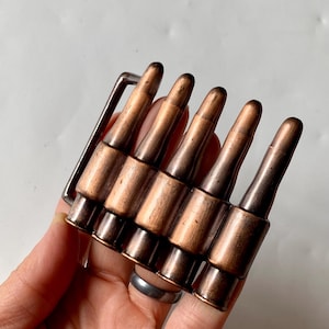 以下が含まれることがあります： 5つの薬莢が付いた銅色のベルトバックル。薬莢は一列に並んでおり、銀色の長方形のバックルに取り付けられています。金属仕上げは、バックルに頑丈で工業的な美しさをもたらします。