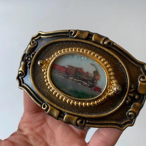 以下が含まれることがあります： 赤と黒の蒸気機関車のミニチュア絵画が描かれた、楕円形の中心部を持つアンティーク真鍮色のベルトバックル。バックルは、縁に装飾的なスクロールのようなデザインと、列車の画像の周りにビーズのボーダーが付いています。