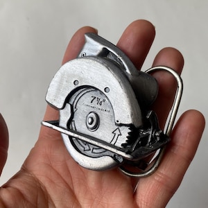 Op de afbeelding: Zilverkleurige metalen riemgesp in de vorm van een cirkelzaag. Het zaagblad is gemarkeerd met "7 1/4" COMBINATION BLADE". De gesp heeft een zilverkleurige metalen lus voor de riem.