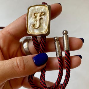 Op de afbeelding: Een vintage bolo stropdas met een rechthoekige hanger. De hanger heeft een parelmoer achtergrond en een goudkleurige letter "F". De hanger is goudkleurig omlijst en bevestigd aan een rood-zwart gevlochten koord met zilverkleurige metalen uiteinden.