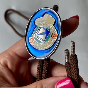 May include: An oval bolo tie with a silver-colored frame. The center features a design with a cowboy hat, text that reads "CALGARY", and the CUPE logo. The background is blue. The tie has a brown cord.