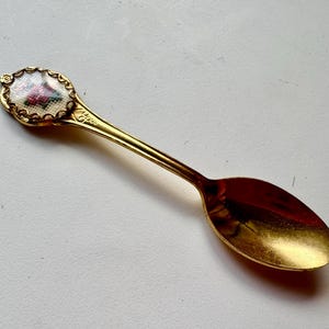 May include: A gold-coloured spoon with a decorative handle. The handle has a small, oval design with a floral pattern in pink, red, and green. The spoon's bowl is a shiny gold colour.