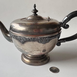 May include: Antique silver-plated teapot with a black handle. The teapot features a decorative floral band around the middle and a small finial on the lid. The spout curves gracefully, and the handle has a curved design. A coin is next to the teapot.
